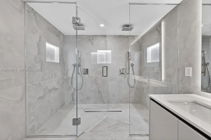 Walk-in shower featuring floor-to-ceiling stone-finish tile, dual ceiling-mounted rainfall showerheads, handheld sprayers, recessed wall niches, and frameless glass enclosure