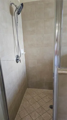 Shower stall featuring neutral-toned wall tiling, diamond-patterned floor tile, a handheld shower fixture, and a glass-enclosed door