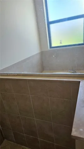 Soaking tub featuring a tiled surround, a neutral-toned stone ledge, and a large window
