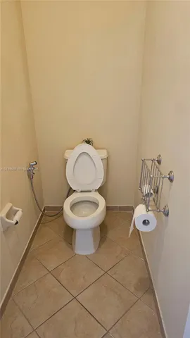 Porcelain toilet with bidet attachment, ceramic tile flooring, and wall-mounted metal toilet paper holder and shelving unit