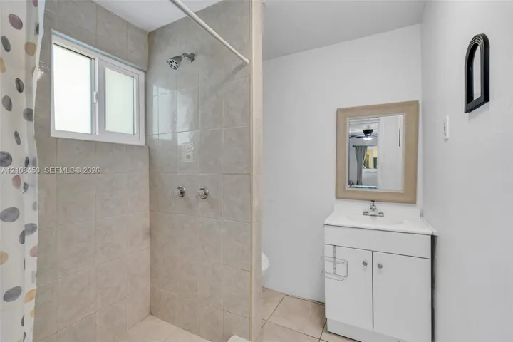 Bathroom featuring tile-surround shower, white vanity cabinet with integrated sink, rectangular wall-mounted mirror, and tile flooring