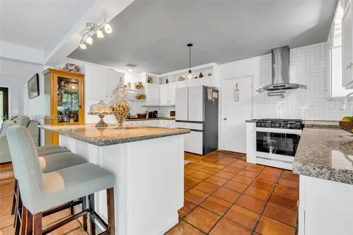Kitchen featuring an island with a stone-finish countertop, white cabinetry, herringbone-patterned wall tile, stainless steel range hood, and terracotta-style flooring