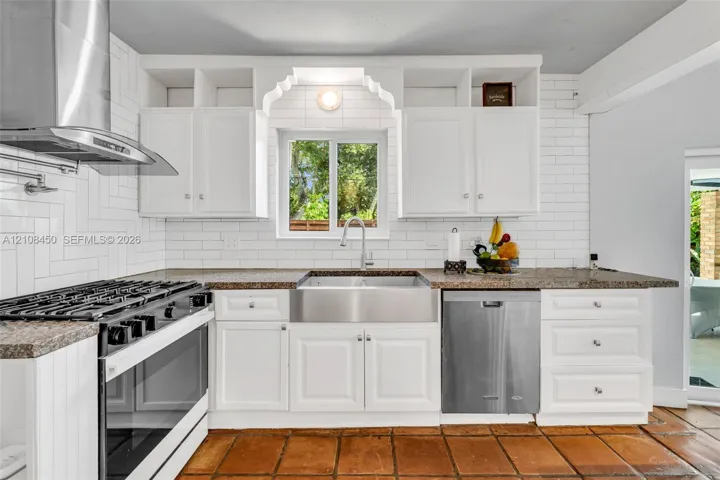 Kitchen featuring custom white cabinetry with decorative arch detailing, stainless steel farmhouse sink, herringbone-patterned subway tile backsplash, stone-surface countertops, and terracotta tile flooring