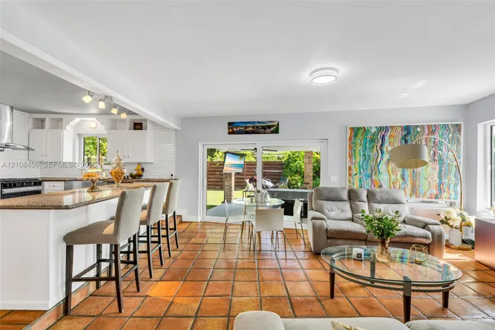 Open-concept kitchen and living area featuring ceramic tile flooring, kitchen island with stone countertop, white shaker-style cabinetry, white tile backsplash, and sliding glass doors