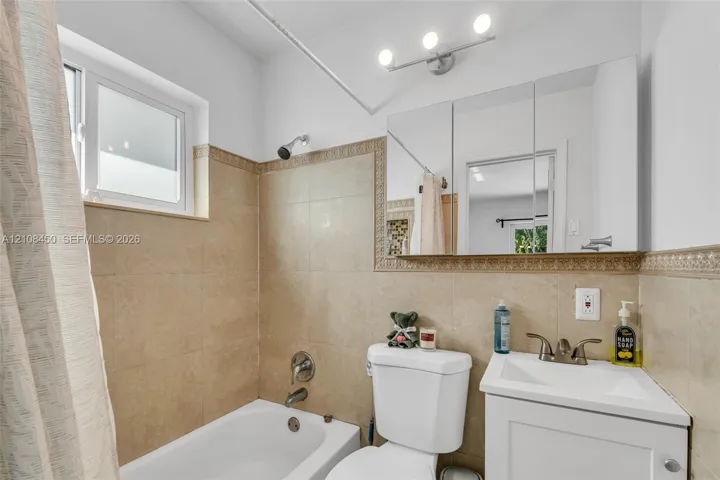 Bathroom featuring a white vanity cabinet, rectangular basin with dual-handle faucet, mirrored medicine cabinet, three-light fixture, and neutral tile surround with decorative border