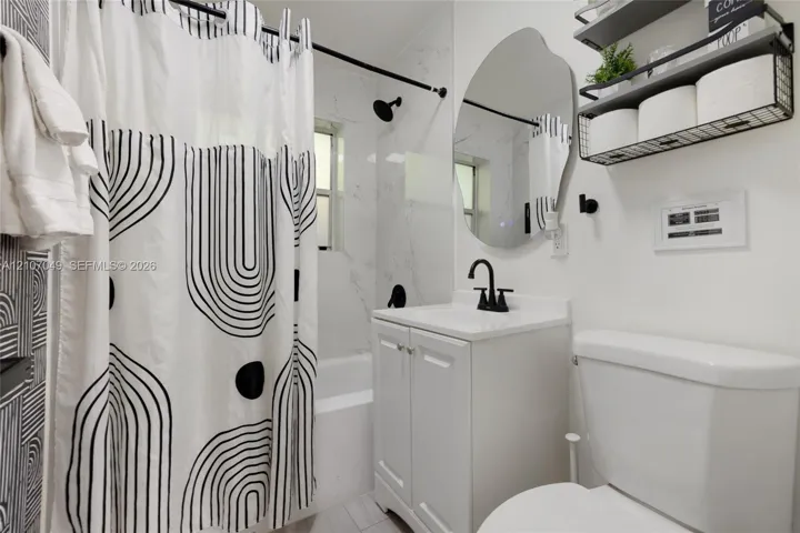 Bathroom featuring a vanity with a white cabinet, stone-finish countertop, and matte black faucet, accompanied by a tub with light-toned wall tile and a matte black shower fixture, complemented by an irregular-shaped wall mirror