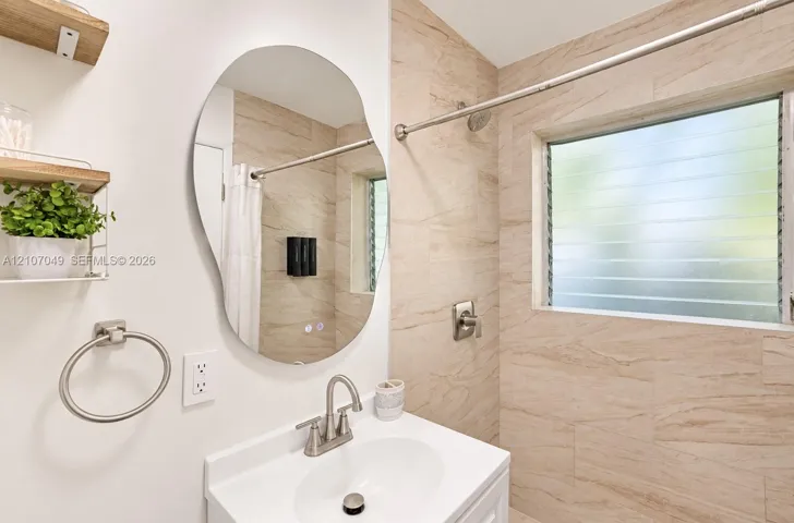 Bathroom featuring a vanity with a white basin, an irregular-shaped wall mirror, louvered glass window, neutral-toned tiled shower surround, and satin nickel fixtures