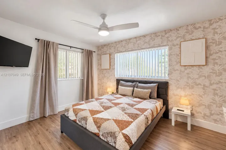 Bedroom featuring wood-finish flooring, patterned wallpaper accent wall, dual windows with vertical blinds, and a central ceiling fan with integrated lighting