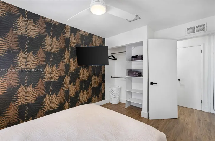 Accent wall with botanical wallpaper, wood-finish flooring, white ceiling fan with integrated lighting, and built-in closet featuring shelving and hanging rods