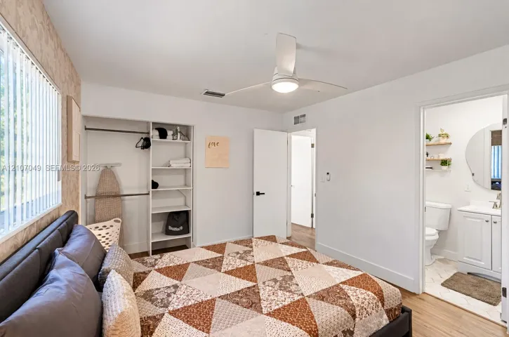 Bedroom featuring wood-finish flooring, a ceiling fan with integrated lighting, a built-in closet system with open shelving, and a bathroom entry with a vanity and wall-mounted circular mirror