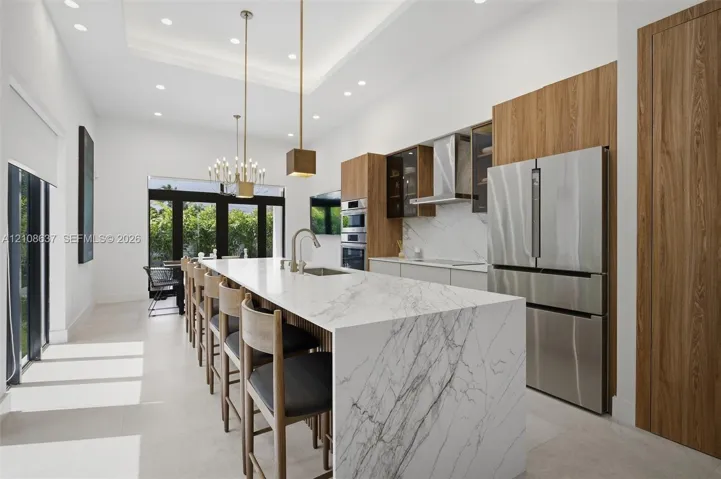 Kitchen featuring a waterfall-edge island with stone-finish countertops, flat-panel wood cabinetry, integrated stainless steel appliances, recessed lighting, and a tray ceiling