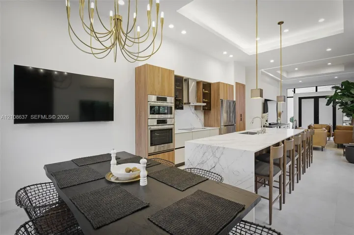 Open-concept kitchen featuring an expansive center island with stone-finish waterfall countertop, integrated stainless steel appliances, flat-panel cabinetry, tray ceiling with recessed lighting, and contemporary metal light fixtures