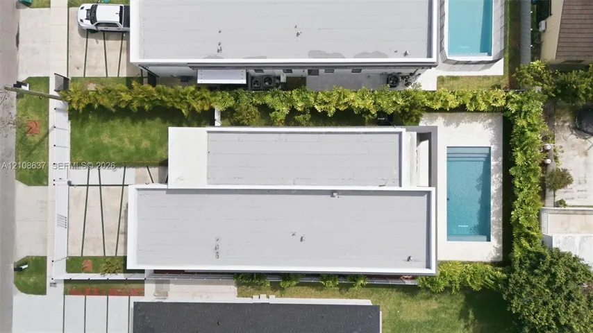 Aerial view of a rectangular swimming pool, manicured lawn area, paved driveway with grass strip accents, and perimeter privacy hedging