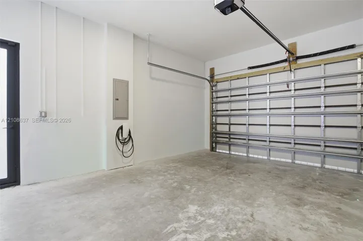Garage featuring concrete flooring, a wall-mounted electric vehicle charging station, an integrated electrical panel, and a metal roll-up door mechanism