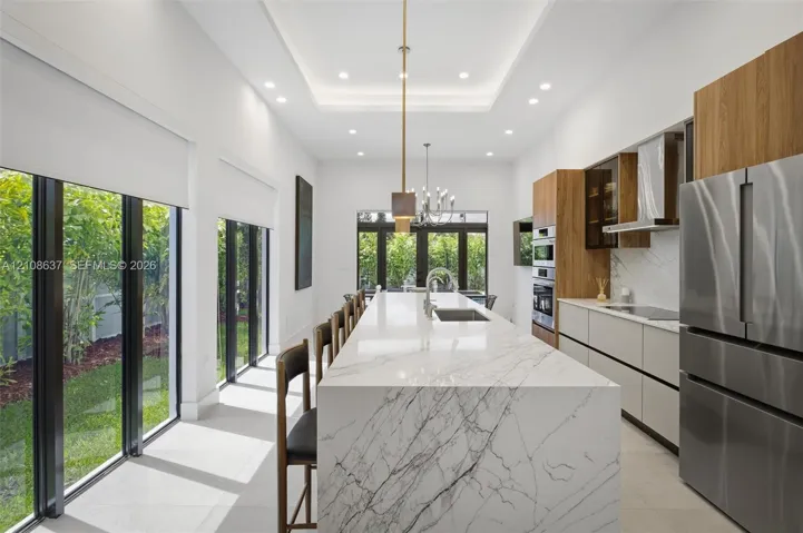 Kitchen featuring a waterfall-edge island with stone-finish surfaces, custom wood-finish cabinetry, built-in stainless steel appliances, recessed ceiling lighting, and floor-to-ceiling windows