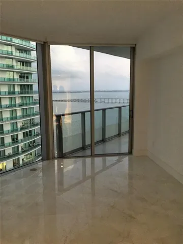 Floor-to-ceiling glass sliding doors, stone-finish flooring, and a private balcony featuring a glass railing and water backdrop