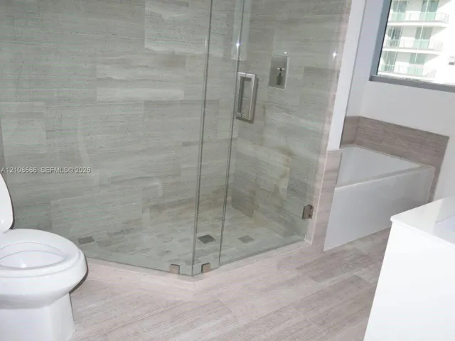 Walk-in glass-enclosed shower featuring large-format stone-finish tile surround and floor, deep soaking tub with tile surround, and neutral wood-finish flooring