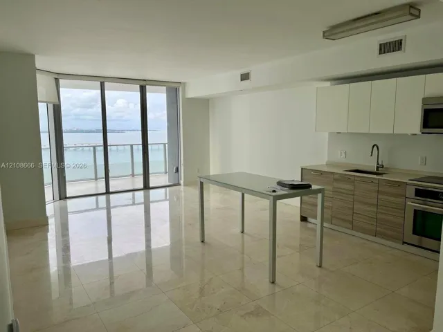 Open-concept kitchen featuring two-tone cabinetry, stone-finish countertops, stainless steel appliances, polished stone flooring, and expansive floor-to-ceiling glass panel doors
