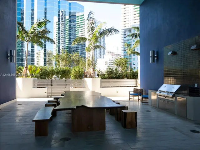 Covered outdoor terrace featuring a built-in stainless steel grill, stone tile flooring, wall-mounted cylinder sconces, and a tile backsplash