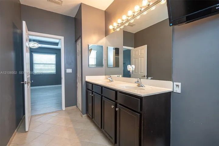 Dual-sink vanity with a solid-surface countertop, dark cabinetry, full-width mirror, and multi-bulb Hollywood-style lighting, complemented by tile flooring