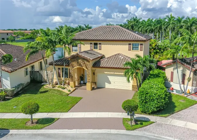 Two-story stucco exterior with stone veneer accents, multi-faceted clay tile roof, paved driveway, arched entryway, and mature palm tree landscaping