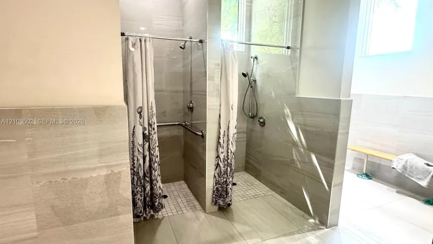 Dual walk-in shower stalls featuring large-format neutral wall tiling, mosaic floor tile, chrome shower fixtures with handheld sprayers, and a built-in wall bench