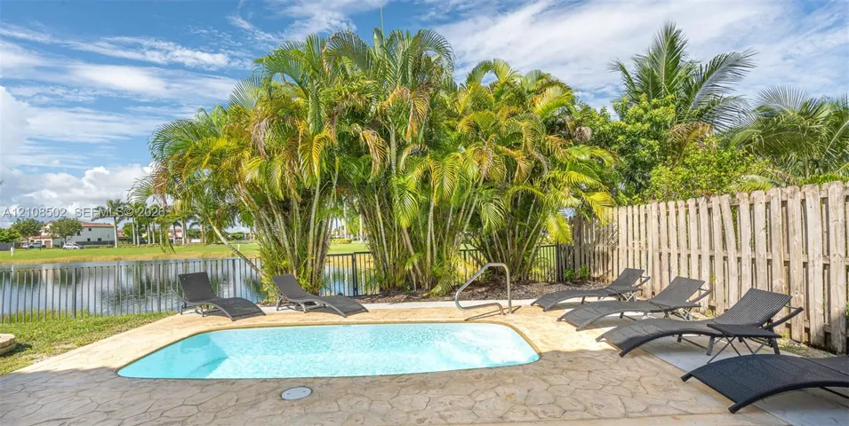 In-ground swimming pool with stone-finish decking, black metal perimeter fencing, and mature tropical landscaping