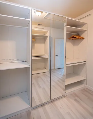 Custom wardrobe cabinetry featuring white shelving, integrated clothing rods, mirrored sliding doors, and wood-finish flooring