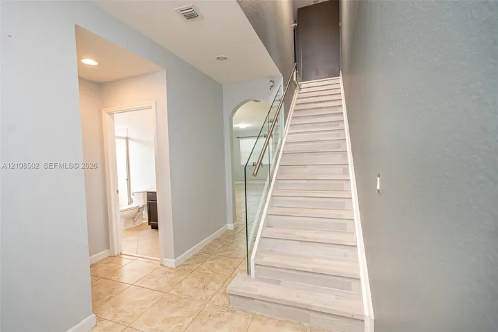 Staircase featuring wood-finish treads and a glass railing with a metal handrail, complemented by large-format tile flooring and an arched entryway