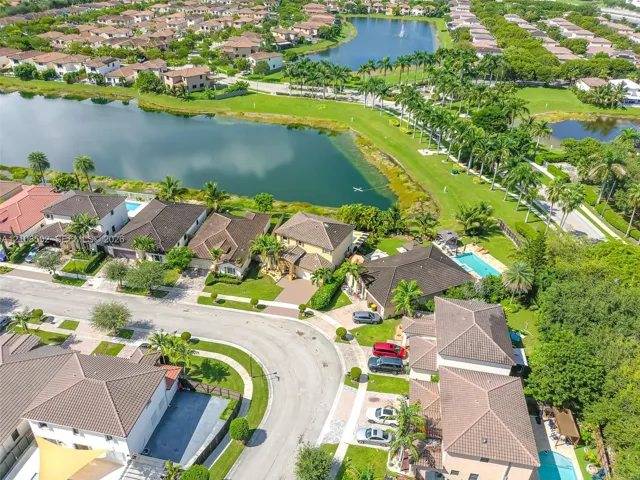 Aerial perspective featuring a multi-parcel residential community with winding paved streets, manicured green space, mature landscaping, and a central lake