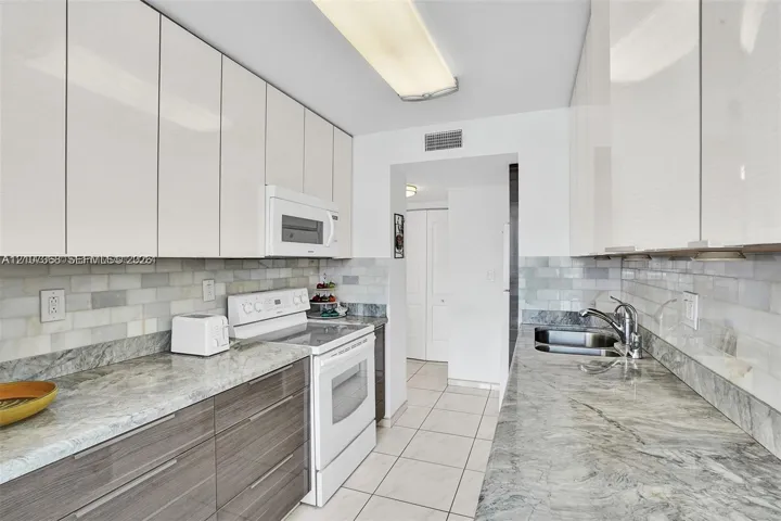 Kitchen featuring high-gloss white upper cabinetry, horizontal grain wood-finish lower cabinets, stone countertops, a subway tile backsplash, and ceramic floor tiling