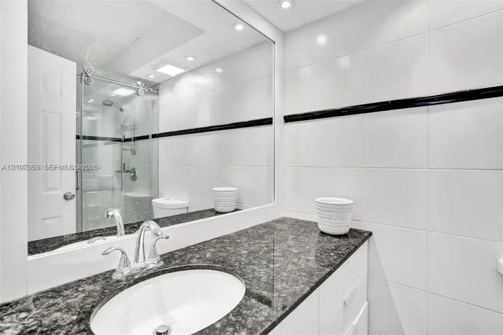 Bathroom featuring a vanity with stone-finish countertop, under-mount sink, polished chrome faucet, full-width mirror, and a glass-enclosed shower with white wall tiling accented by a horizontal dark tile border
