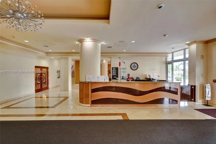 Lobby featuring a polished stone-tile floor with decorative inlay, a custom curved reception desk with wood-tone wave detailing, structural columns, and a large multi-point contemporary chandelier