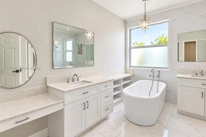 Freestanding soaking tub with chrome wall-mounted faucet, dual white vanities with stone countertops, integrated open shelving, and large-format tile flooring