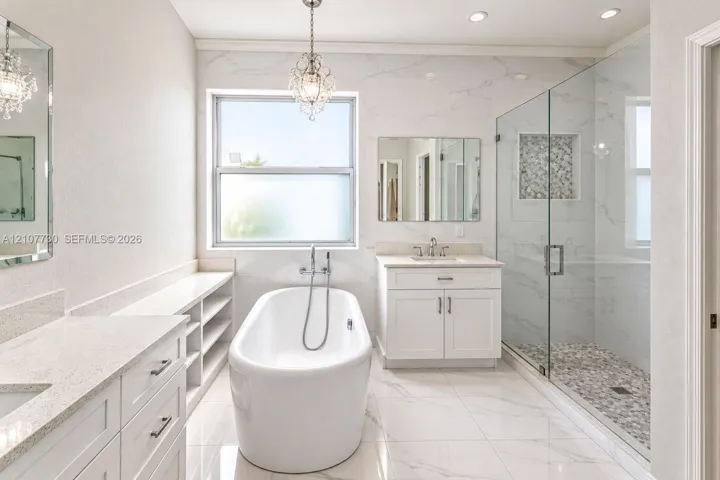 Bathroom featuring a freestanding soaking tub, walk-in glass-enclosed shower with a stone-tile floor, dual white vanities with solid-surface countertops, and decorative pendant lighting