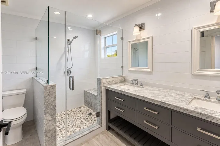 Double vanity with stone countertop, glass-enclosed walk-in shower featuring an integrated bench seat, pebble-style shower flooring, and wall-mounted light fixtures