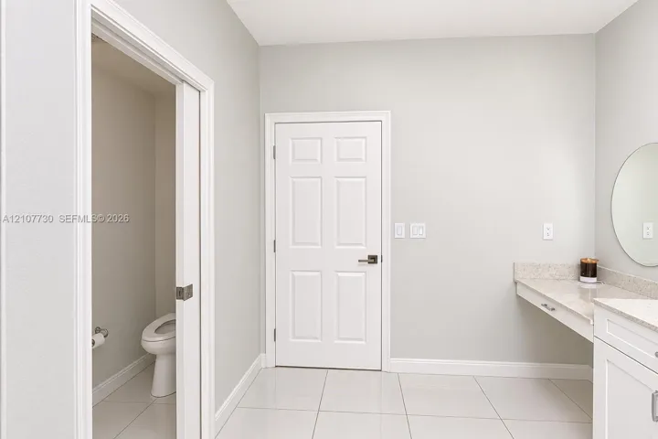 Light-toned tile flooring, white six-panel door, built-in vanity with stone-composite countertop, and wall-mounted circular mirror