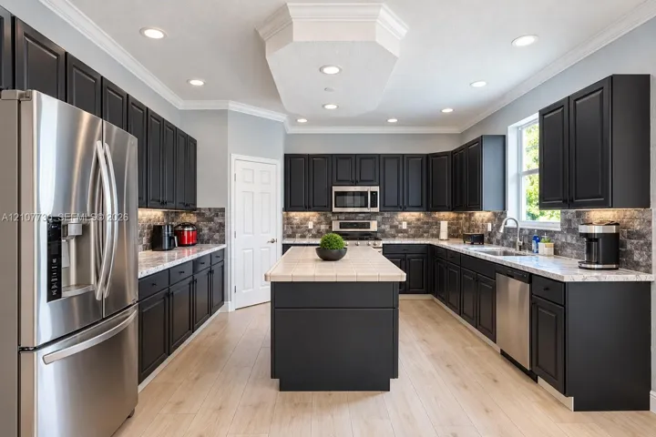 Kitchen featuring dark-toned cabinetry, light-toned countertops, tile-topped center island, stone-finish backsplash, and stainless steel appliances