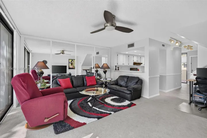 Living area featuring a ceiling fan, wall-to-wall mirrored panels, carpeted flooring, and a pass-through opening to the kitchen