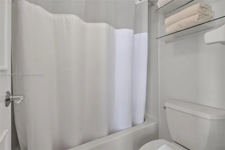Bathroom featuring a white bathtub, tiled shower surround, dual glass shelving unit, and standard toilet