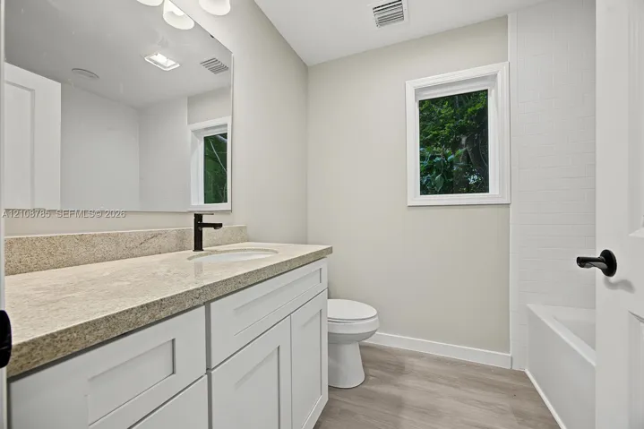 Bathroom featuring a white shaker-style vanity, stone-surface countertop, dark metal hardware, wood-finish flooring, and a subway-tile tub surround