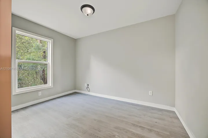 Neutral-toned interior space featuring wood-finish flooring, a single-hung window with exterior greenery, and a flush-mount ceiling light fixture
