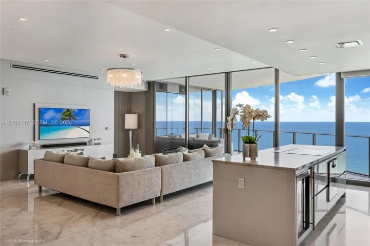Open-concept interior featuring polished stone flooring, recessed lighting, a modern crystal chandelier, floor-to-ceiling glass panel walls, and a kitchen island with a stone countertop