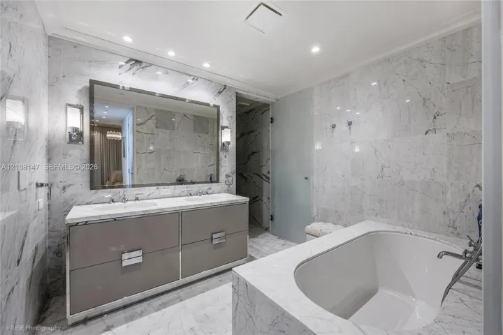 Dual-sink vanity with high-gloss cabinetry, stone-surround soaking tub, wall-to-wall stone tiling, and recessed lighting