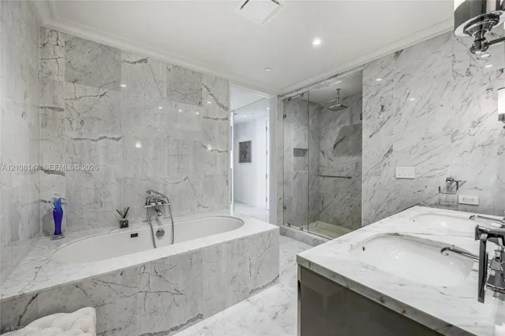 Bathroom featuring floor-to-ceiling stone tile surround, soaking tub with wall-mounted hardware, glass-enclosed walk-in shower, and dual-basin vanity with stone countertops