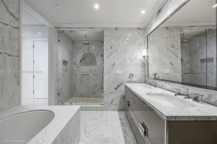 Bathroom featuring extensive stone-surround, frameless glass-enclosed shower with built-in recessed shelving, integrated soaking tub, custom dual-sink vanity with stone countertop, and wall-to-wall mirror