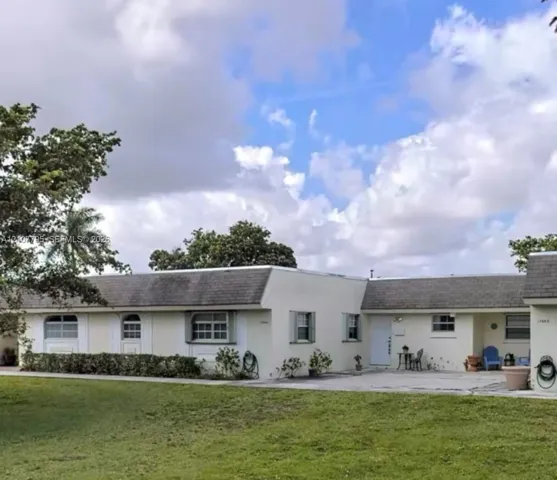 Single-story exterior featuring a light-colored stucco facade, shingled roof, concrete patio, and decorative window awnings