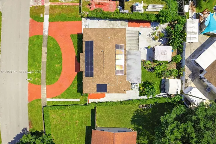 Aerial perspective featuring a residential lot with solar panel arrays, a multi-section roof, a paved concrete driveway, and mature landscaping