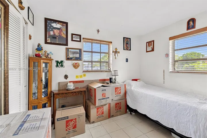 Room featuring neutral-toned tile flooring, dual-aspect windows with horizontal blinds, and a floor-to-ceiling built-in corner cabinet with glass-panel doors