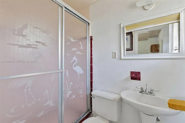 Bathroom featuring a semi-frameless sliding glass shower enclosure with etched decorative detailing, a white pedestal sink with dual-handle chrome faucet, a rectangular wall-mounted mirror with integrated lighting, and a built-in recessed soap dish